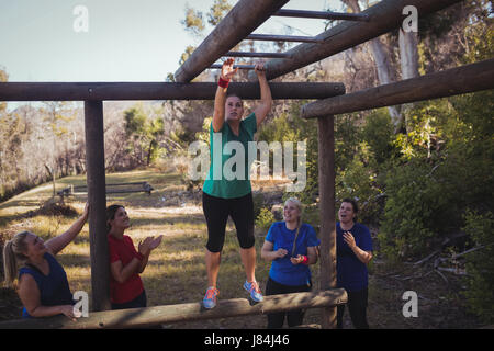 Woman being cheered bye her teammates to climb monkey bars during obstacle course training in the boot camp Stock Photo