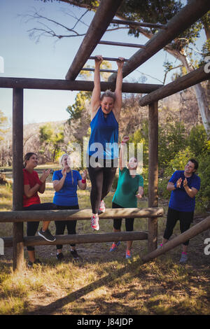 Woman being cheered bye her teammates to climb monkey bars during obstacle course training in the boot camp Stock Photo