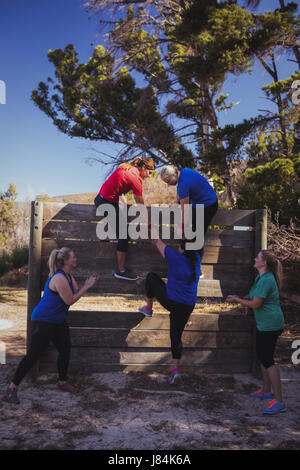 Woman being assisted by her teammates to climb a wooden wall during obstacle course training at boot camp Stock Photo