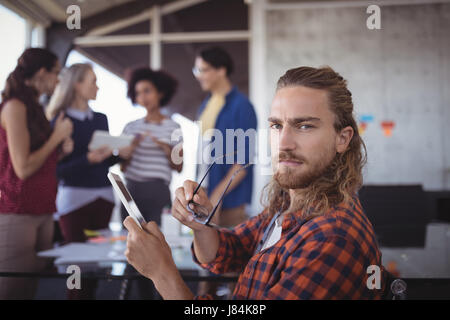 Portrait of serious businessman holding eyeglasses and digital tablet with colleagues in background Stock Photo