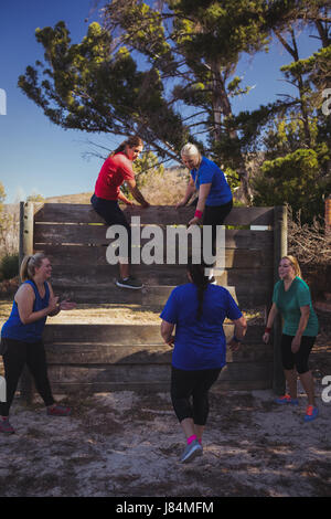 Woman being cheered by her teammates to climb a wooden wall during obstacle course training at boot camp Stock Photo
