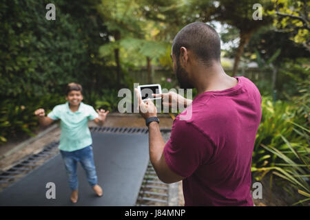 Rear view of father photographing son jumping on trampoline at park Stock Photo