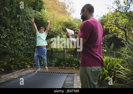 Rear view of father photographing son while jumping on trampoline at park Stock Photo