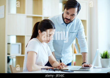 Portrait of Asian man helping young woman working with laptop in office, giving instructions for project Stock Photo
