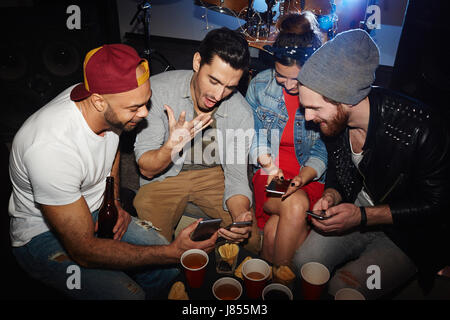 Friendly guys and girl reading messages in smartphone during get-together Stock Photo