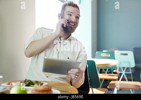 Portrait of modern handsome man in casual shirt speaking by smartphone and using digital tablet while having lunch in cafe Stock Photo