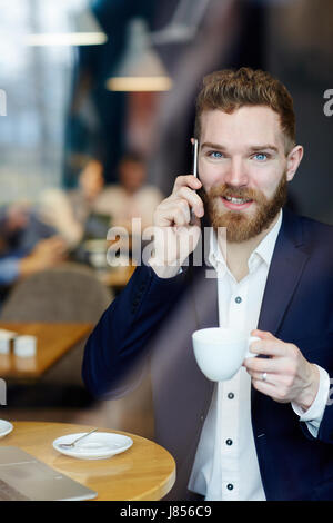 Portrait of handsome young businessman speaking by phone in cafe and looking at camera smiling charmingly Stock Photo