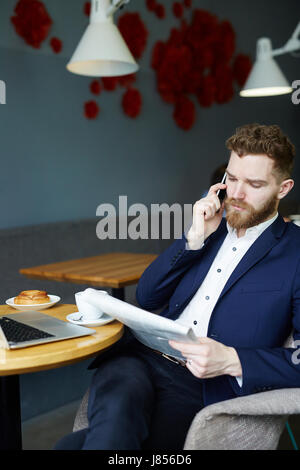 Portrait of handsome young businessman speaking by smartphone while reading morning newspaper during breakfast  in cafe Stock Photo