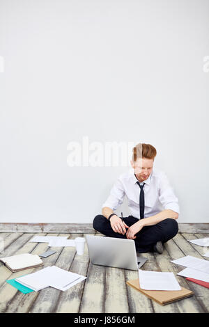 Portrait of modern young businessman sitting cross legged on floor in office thinking while using laptop with documents around Stock Photo