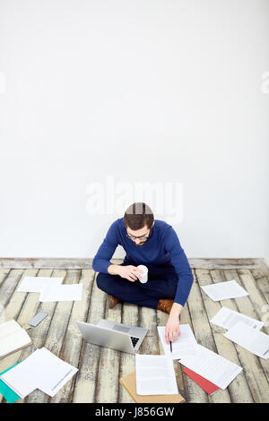 Portrait of  modern freelancer sitting cross legged on floor in office,  working with laptop and  documents laid out around Stock Photo