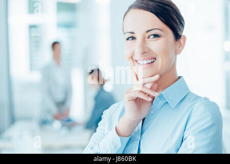 Successful businesswoman posing in her office and smiling at camera, office workers on background, selective focus Stock Photo