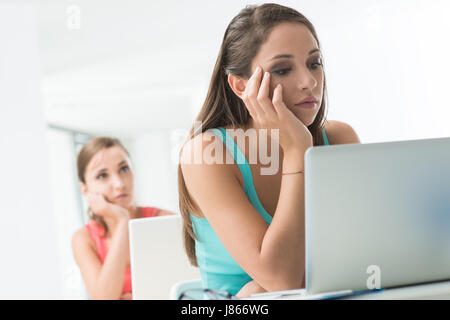 Bored tired girl sitting at school desk with hand on chin and using a laptop Stock Photo