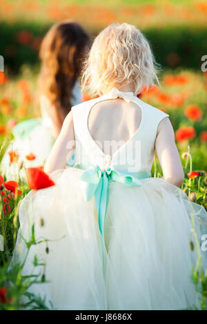 childcare, freedom, countryside, fashion, style concept - sorrounded by tall poppy flowers two fair and dark haired girls, wearing sunday outfits, walking through the field Stock Photo