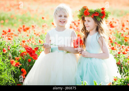 freedom, celebration, dream, happy childhood concept - in the poppy field two giggling girlfriends in white and blue sleeveless dresses holding posies of wild flowers Stock Photo