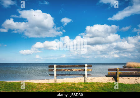 Bench on the sandy beach at the sea on the background of colorful blue sky with clouds at sunset. Beautiful landscape with empty old wooden bench stan Stock Photo