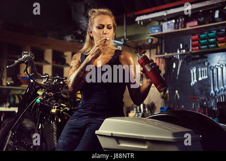 Woman mechanic lights up a cigarette with a gas burner in a motorcycle workshop Stock Photo
