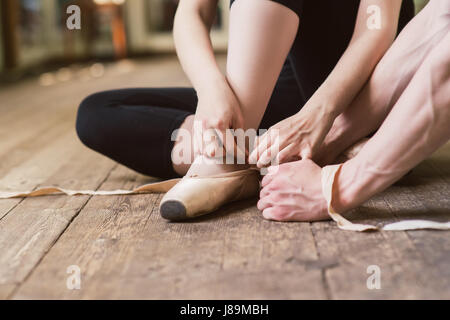 Young ballerina or dancer girl putting on her ballet shoes on the wooden floor. Male ballet dancer helps puttiing on. Ballet dancer tying ballet shoes