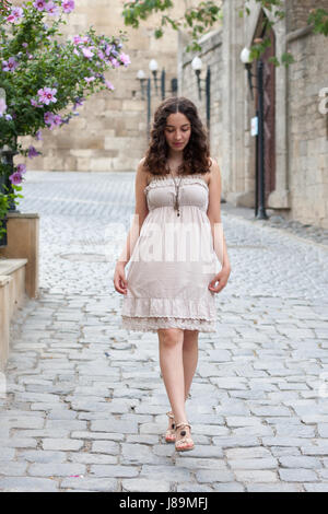 Full length portrait of curlie young girl wearing biege romantic dress strolling through old streets of Inner City of Baku looking down and holding dr Stock Photo