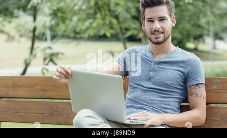 Young handsome man relaxing at the park, sitting on a wooden bench and using a laptop Stock Photo