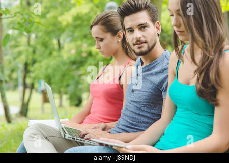 Adolescents at the park relaxing, one is reading a book, a guy is using a laptop and a girl is using a digital tablet Stock Photo