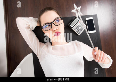 young business woman with moody expression on her face laying at the table on her working place at office and holding magic wand in her hand. top view. Stock Photo