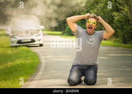 Young man on his knees on the road. He is under stress and pulling his hair out because his car is broken and in smoke. Travel and vacation road probl Stock Photo