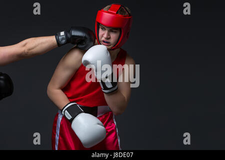 Teenage boxer in red form and helmet is being punched into face. Studio shot on black background. Copy space. Stock Photo
