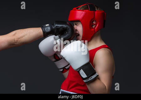 Teenage boxer in red form and helmet is being punched into face. Studio shot on black background. Copy space. Stock Photo