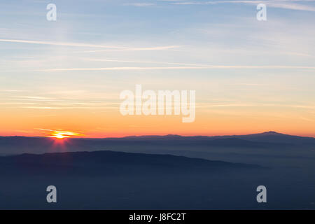 Sun coming down behind some misty mountains, the sky is orange and blue, with many long and thin clouds and jet trails Stock Photo