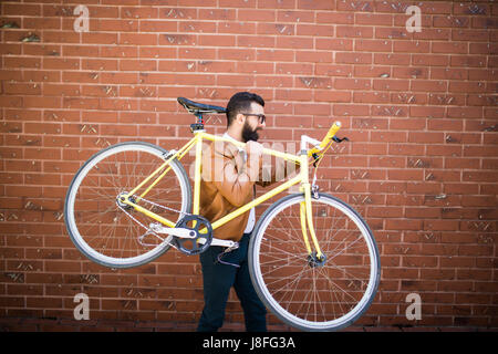 Bearded man with a fixie bicycle. Confident bearded man carrying his bicycle on shoulder and looking at camera while walking outdoors. Stock Photo