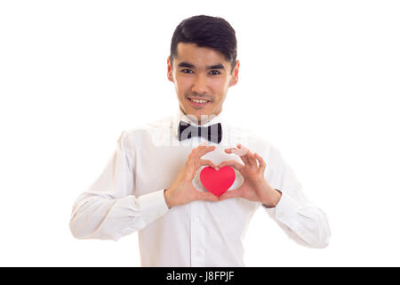 Pleasant man with black hair in white T-shirt with black bow-tie holding a red paper heart on white background in studio Stock Photo