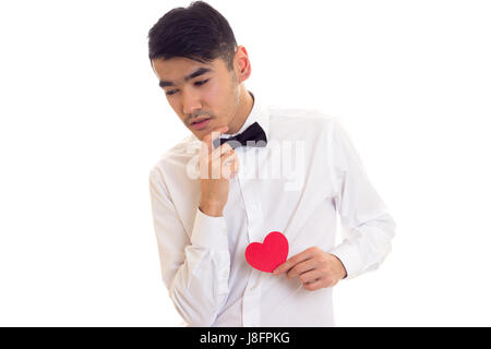 Handsome young man with dark hair in white T-shirt with bow-tie holding a red paper heart on white background in studio Stock Photo