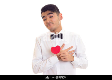 Attractive young man with dark hair in white T-shirt with bow-tie holding a red paper heart on white background in studio Stock Photo
