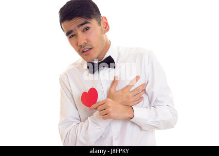 Confident young man with dark hair in white T-shirt with bow-tie holding a red paper heart on white background in studio Stock Photo