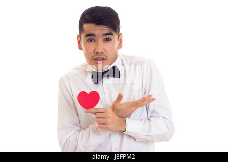 Nice young man with dark hair in white T-shirt with bow-tie holding a red paper heart on white background in studio Stock Photo
