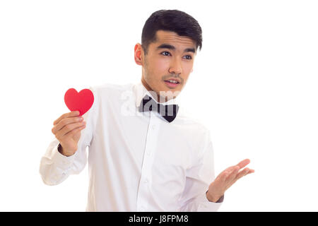 Pleasant young man with dark hair in white T-shirt with bow-tie holding a red paper heart on white background in studio Stock Photo