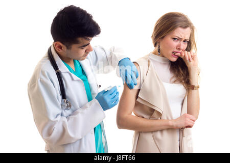 Young smartlooking doctor with dark hair in white gown with stethoscope and blue gloves making an injection of young scared woman with long brown hair Stock Photo