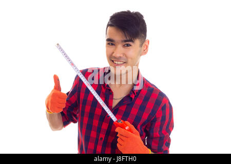 Young positive man with dark hair in red plaid shirt with orange gloves holding a tape-measure on white background in studio Stock Photo