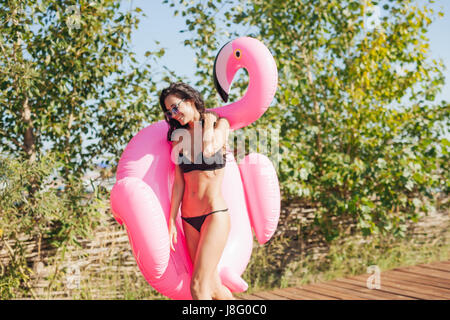 Cheerful beautiful young millennial woman holding sprinkled pink inflatable flamingo float in summer. 20s Stock Photo