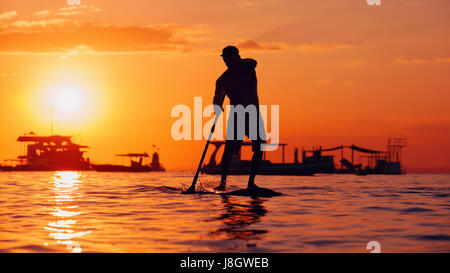 Active paddle boarder. Black sunset silhouette of young sportsman paddling on stand up paddleboard. Healthy lifestyle. Water sport, SUP surfing tour Stock Photo