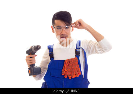 Handsome young man with dark hair in white shirt and blue overall with orange gloves and protective glasses holding electric screwdriver on white back Stock Photo