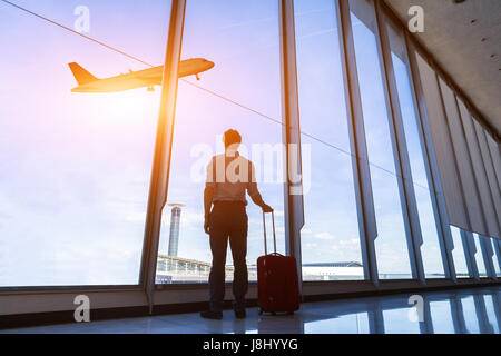 Businessman with suitcase at airport international departure gate looking at flying airplane through the windows Stock Photo