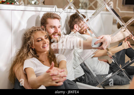 Smiled couple making a selfie sitting on the floor near the table. Blonde man and woman in white T-shirts. Stock Photo