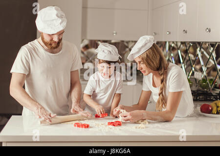 Happy family of father, mother and little son making dough together in the modern white kitchen. Family wearing white aprons and hats. Stock Photo