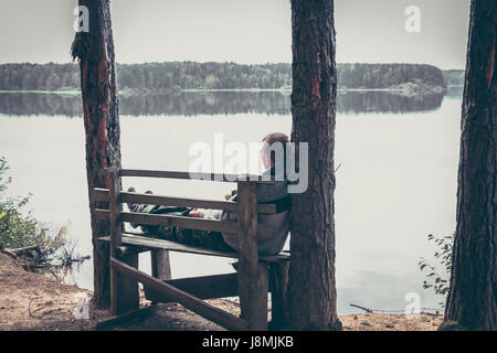 Pensive man on the high edge of river bank sitting on the bench and looking on beautiful scenery with tranquil water Stock Photo