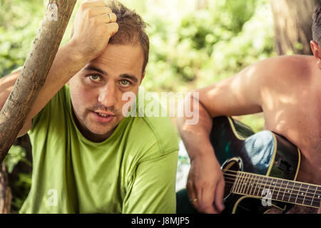 Candid young man with sincere look singing with guitar during outdoor camping party among foliage Stock Photo