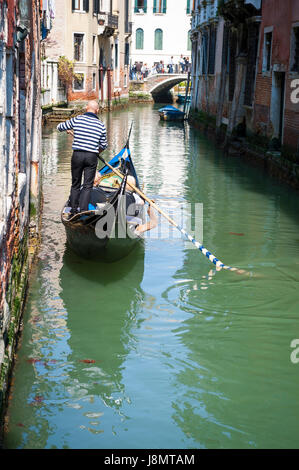 Scenic view of a small canal in Venice, Italy, with gondolier in traditional striped shirt maneuvering gondola along green waters Stock Photo