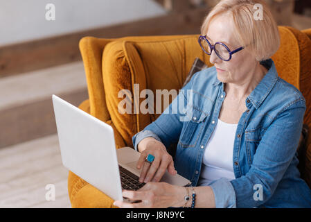 I will check. Confident lady wearing round glasses sitting in yellow and soft armchair while using her device Stock Photo