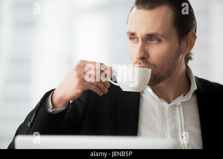 Portrait of young man drinking coffee and looking toward window. Businessman takes coffee-break at workplace. Thoughtful office worker holding white p Stock Photo