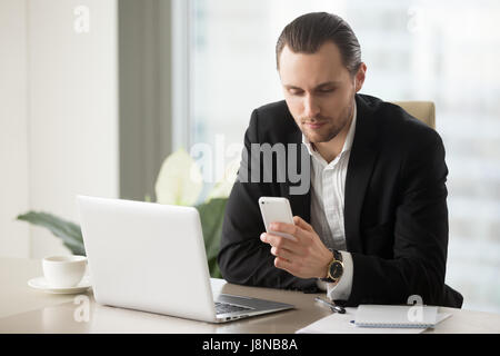 Successful businessman dials number on cellphone at desk with laptop. Attractive entrepreneur using mobile app on phone, guy reading message or news,  Stock Photo